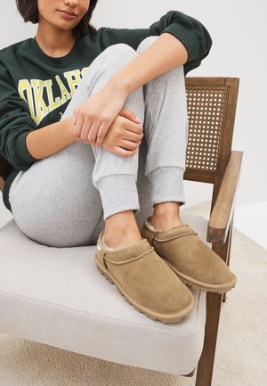 Zoccoli in pelle scamosciata marrone con profilo basso e rivestimento morbido, posati su una sedia imbottita di colore grigio chiaro accanto a pantaloni della tuta grigi.