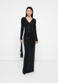 Dlouhé černé šaty se štrasovými flitry, s výstřihem do V, dlouhými rukávy a slim fit střihem. Hladký povrch s jemným reflexním vzorem. Pár černých podpatků a kabelka.