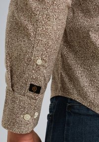 Braun gemustertes Hemd mit beigem Hintergrund, mit einem geknöpften Manschette und einem Markenetikett; kombiniert mit dunkelblauen Denimhemd.