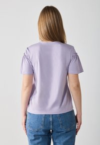 T-shirt lilla a maniche corte con dettagli arricciati sulle spalle, collo rotondo e vestibilità comoda, abbinato a jeans in denim blu a vita alta.