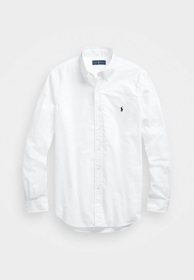 Bela dolg rokav srajca s gumbi iz bombaža. Ima klasičen ovratnik, gumbnice na zapestjih in majhen temno moder vezen logotip na prsih.