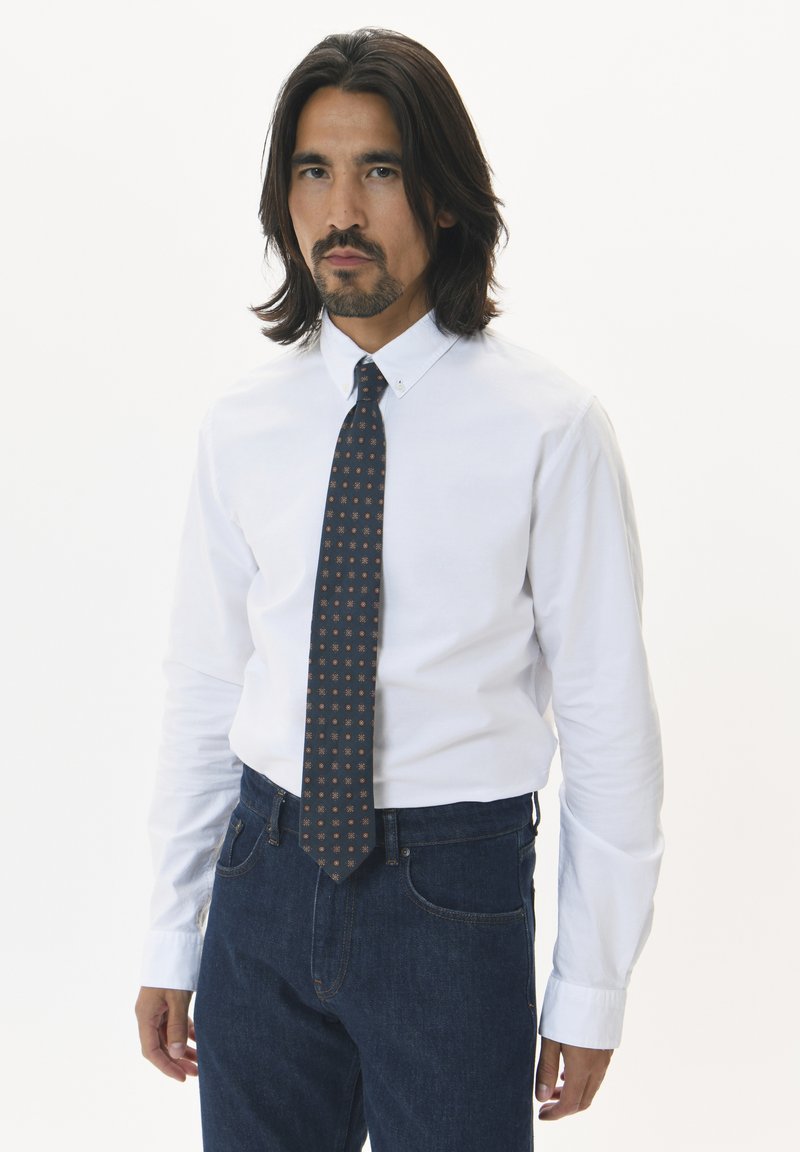 Vit långärmad skjorta med krage som knäpps, kombinerad med mörkblå denimjeans. En mönstrad mörk slips fulländar klädseln.