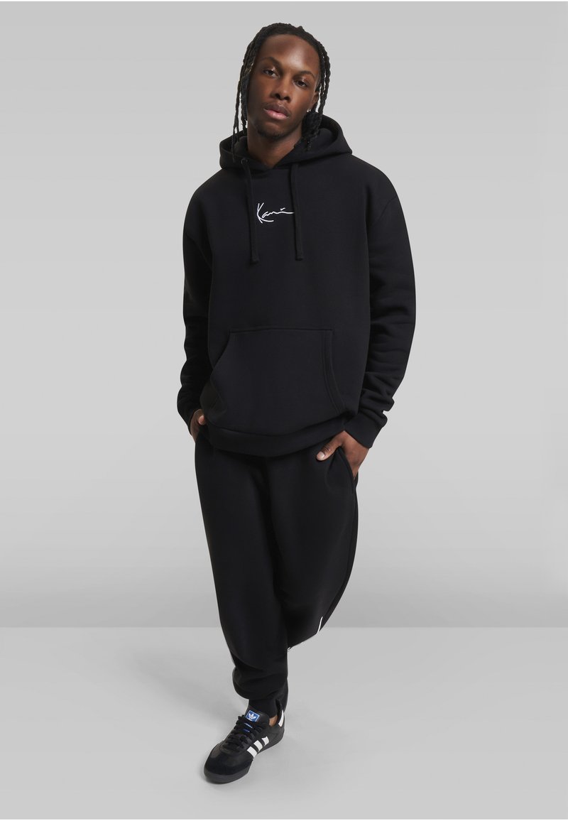 Tânăr cu părul împletit purtând un hanorac negru, pantaloni sport negri și adidași negri Adidas, stând în fața unui fundal gri simplu.