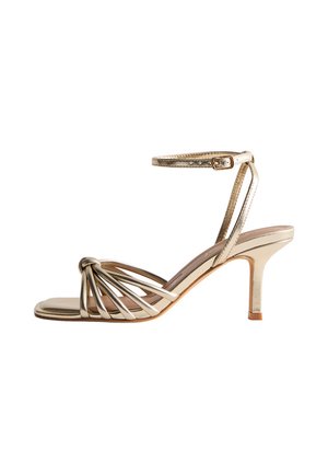 Sandalo strappy metallico dorato con fibbia alla caviglia, dettaglio anteriore annodato, punta aperta e tacco sottile di altezza media su uno sfondo bianco.