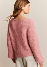 Kvinde med langt hår iført en rosa struktureret striktrøje og lyse jeans, som står sidelæns mod en neutral baggrund.