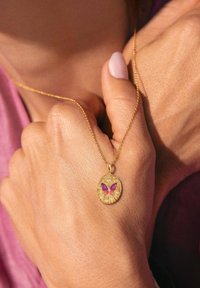 Gouden ketting met een ronde hanger met een paarse vlinderontwerp en decoratieve accenten, vastgehouden tegen een hand met een gestructureerde huid.