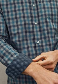 Camisa a cuadros en azul y gris con cuello abotonado, que presenta puños de patrón oscuro y pequeños puntos blancos. Los botones son blancos y redondos.