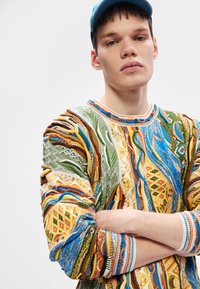 Nuori mies ristittyin käsin, päällään monivärinen teksturoitu neulepaita ja sininen hattu, vaaleaa taustaa vasten.