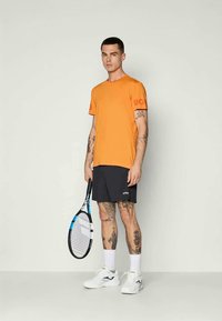 Oranžinė sportinė marškinė su raudonu logotipu ant rankovės, derinama su juodais šortais. Laikomos baltos sportinės bateliai ir teniso raketė.