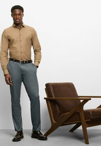 Hellbraunes Hemd mit Knöpfen, graue maßgeschneiderte Hose, schwarze Lederschuhe und ein brauner gepolsterter Stuhl mit Holzrahmen. Einfaches, modernes Design.