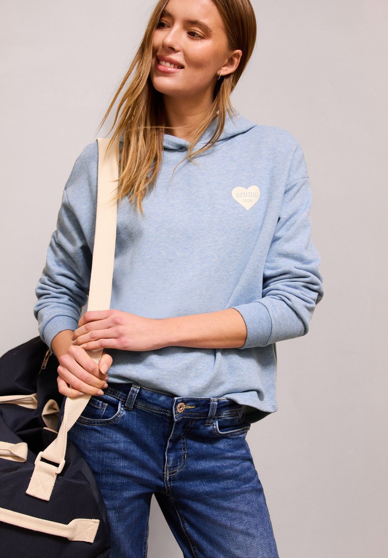 Jeune femme portant un sweat à capuche bleu clair et un jean, transportant un sac de sport noir avec des sangles beige, souriant et regardant sur le côté devant un fond uni.