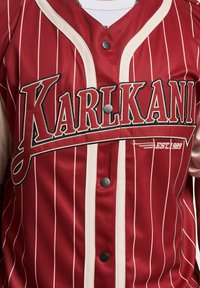 Piros hálós baseball mez függőleges fehér csíkokkal, fekete gombokkal és hímzett "KARL KANI EST. 1989" logóval a mellrészen.