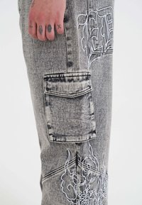 Szare dżinsowe spodnie o teksturowanym wykończeniu, z boczną kieszenią i białymi haftowanymi wzorami, w tym płomieniami i literami.