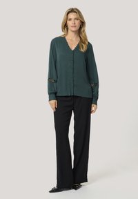 Blusa verde scuro con scollatura a V, chiusura con bottoni e dettagli in pizzo sui polsini, abbinata a pantaloni neri ampi e ballerine nere.
