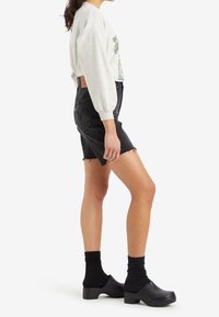 Grå sweatshirt med grafiskt tryck, svart denimshorts med hög midja och fransat avslut, samt svarta träskor som bärs med svarta strumpor.