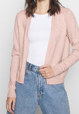 Ljusrosa cardigan i mjukt material, med avslappnad silhuett, öppen framtill, ribbade ärmar och fåll, bärs över en vit topp med blå jeans.