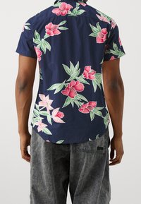 Short-sleeved navy shirt with pink floral pattern and green leaves. Features a relaxed fit and curved hem. Cotton material.