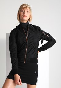 Veste bombardier noire en nylon brillant, avec fermeture à glissière, poignets à côtes et une poche zippée supplémentaire sur la manche.