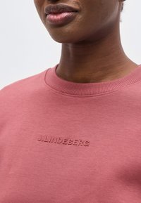 Κοντινό πλάνο ενός ατόμου που φοράει φροντισμένη ροζ φούτερ μπλούζα με στρογγυλή λαιμόκοψη και ανάγλυφο λογότυπο «J.LINDEBERG» στο στήθος.