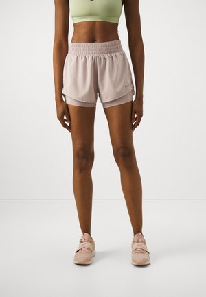 Frau steht, trägt hellgrünen Sport-BH, beige geschichtete Laufshorts und beige Sportschuhe vor weißem Hintergrund.