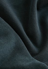 Tume roheline kangas pehme, tekstureeritud pinnaga, millel on peened kudumismustrid ja õrnad voltid, luues sujuva ja voolava välimuse.