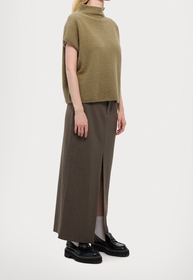 Femme portant un pull à col roulé manches courtes vert olive, une longue jupe marron avec une fente devant, des chaussettes blanches et des mocassins noirs sur un fond neutre.