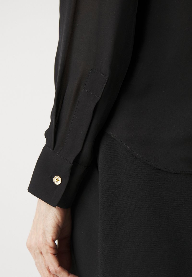 Czarna Bluzka z przezroczystego materiału z długimi rękawami, posiadająca zaokrąglony mankiet z detalem złotego guzika. Gładka tekstura, prosty design.