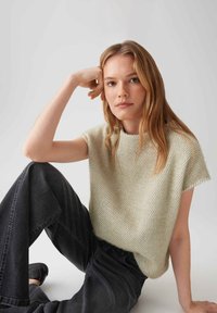 Femme jeune aux longs cheveux blonds, portant un pull crème sans manches à texture et un jean noir, assise avec un bras reposant sur son genou.