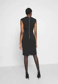 Svart ärmlös klänning med en figurnära design, försedd med ett vertikalt dragkedja i ryggen och en knälång fåll. Slät tyg med en lätt glans.