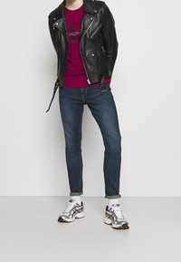 Zwartleren motorjack, bordeauxrood t-shirt met logo, donkerblauwe slim-fit jeans, wit en zilver sportieve schoenen, opgerolde mouwen van de jeans.