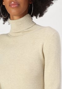 Beige ribbestrikket turtleneck genser med en tettsittende design og myk tekstur, med høy hals og lange ermer.