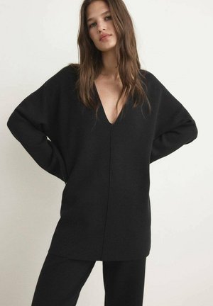 Zwarte oversized trui met een diepe V-hals, lange mouwen en een gladde textuur. Gecombineerd met zwarte broeken, wat een monochromatische look creëert.