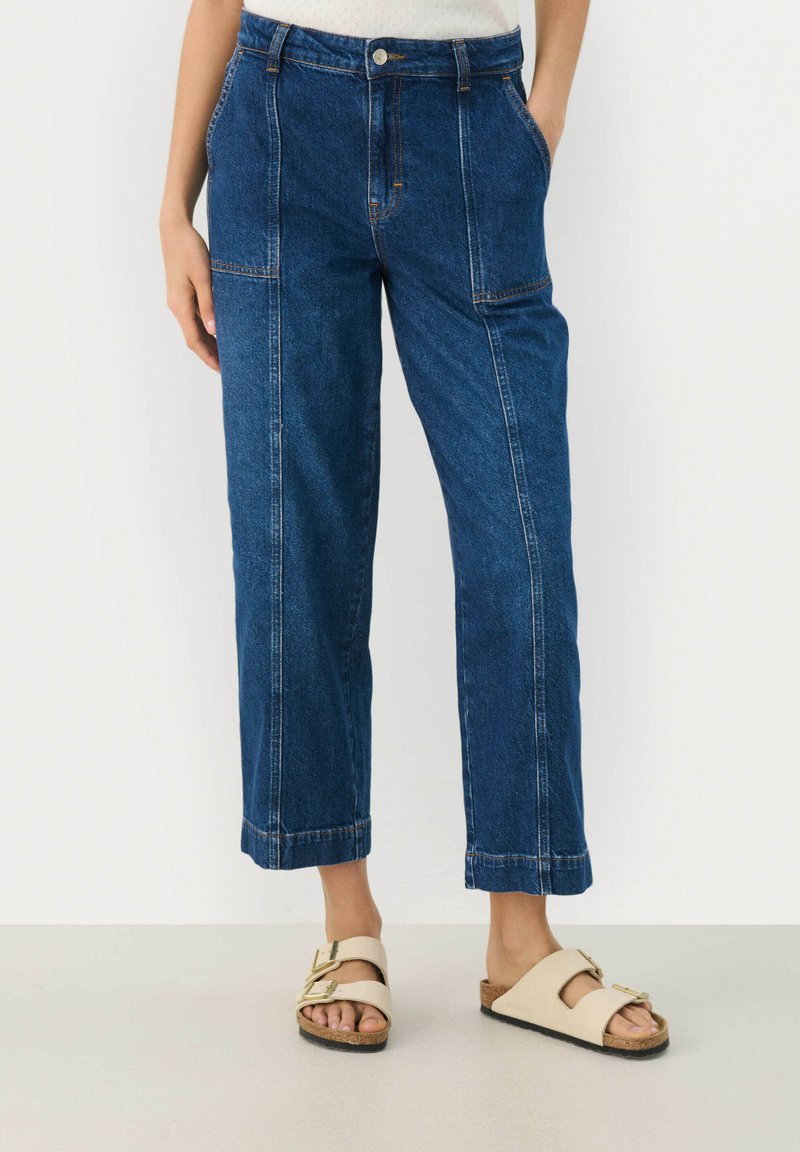 Jeans med vida ben i mörkblått med kontrasterande sömmar, framfickor och kortade avslut. Matchas med beiga sandaler med öppen tå.