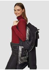Zwarte jas met glanzende zilveren voering, over de schouder gedragen; rode lange mouwen top, donkere jeans; binnenzak met zichtbare onderhoudsinstructies.