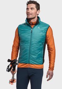 Teeal-farbene isolierte Weste mit hohem Kragen, vorne mit Reißverschluss und schwarzen seitlichen Einsätzen. Getragen über einem orangefarbenen Langarmoberteil, mit Trekkingstöcken.