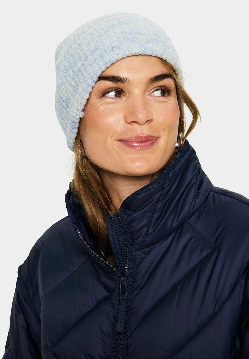 Berretto lavorato a maglia di color blu chiaro con texture a coste, realizzato in materiale morbido. Indossato con un giubbotto isolante di un blu navy scuro con chiusura a zip.