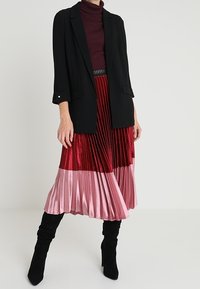 Žena v černém saku, burgundském roláku, plisované midi sukni v červené a růžové barvě a černých kozačkách po kolena, stojící na bílém pozadí.