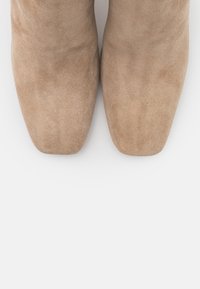 Un par de botas de ante color arena con punta cuadrada se muestran desde arriba sobre un fondo claro.