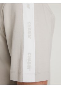 T-shirt grigio chiaro a maniche corte con una striscia bianca e il testo in rilievo "CHASIN'" sulla spalla. Materiale in cotone morbido con cuciture minime.