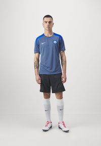 Camiseta deportiva gris con acentos azules, logo en el pecho, combinada con pantalones cortos negros y calcetines blancos. El jugador usa zapatos de fútbol blancos y rojos.