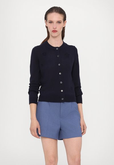 Merino neulecardigan navy-värissä, jossa on pyöreä kaulus, nappikiinnitys ja hienovaraiset tähtikuviot; yhdistetty vaaleansinisii räätälöityihin shortseihin.