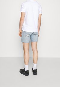 Shorts de mezclilla azul claro con dobladillo desflecado, combinados con una camiseta blanca lisa y zapatillas negras. Detalles sutiles en los bolsillos traseros de los shorts.