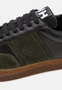 Sneaker in pelle nera con pannelli in suede verde oliva, lacci neri, suola in gomma texturizzata e logo sulla linguetta.