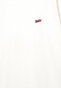 Hvid t-shirt lavet af bomuld, med et lille broderet logo i maroon på venstre brystområde, rund hals design. Glat tekstur.