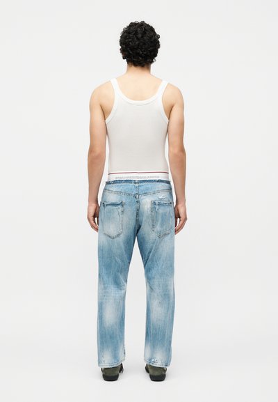 Άνδρας με σγουρά μαλλιά που φοράει λευκή φανέλα, ξεθωριασμένο φαρδύ μπλε τζιν και σκούρα παπούτσια, στεκόμενος με την πλάτη προς απλό φόντο.