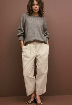 Grauer Pullover mit langen Ärmeln, beigen Faltenhosen und Schuhen mit Leopardenmuster. Glatte Texturen und lockere Passform. Einfarbig brauner Hintergrund.