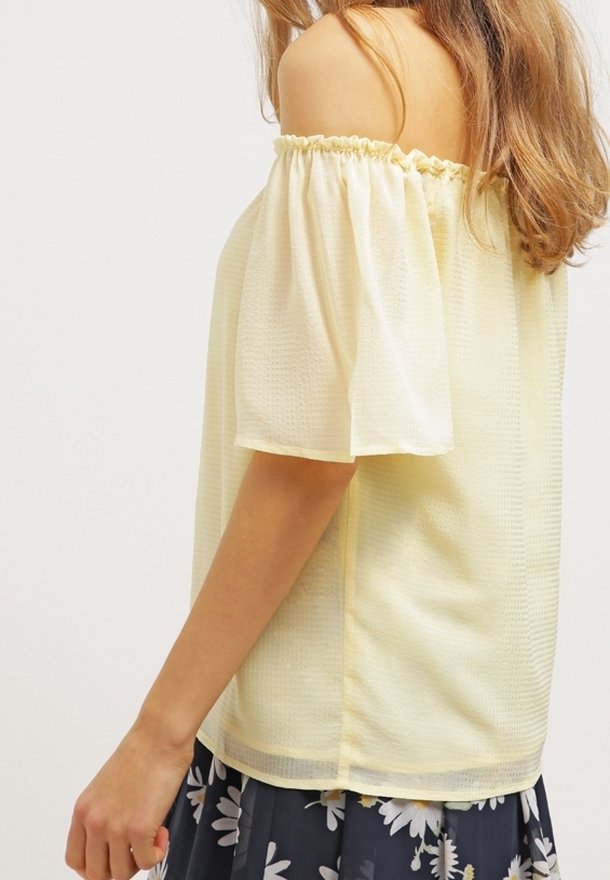 Blouse jaune à épaule dénudée avec un tissu léger et texturé et des manches froncées. La blouse présente une coupe décontractée et un léger brillant.