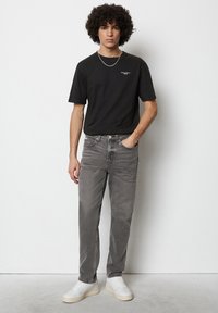 Zwarte katoenen T-shirt met korte mouwen, gecombineerd met grijze tapere jeans en witte sneakers. Krullend haar en een minimale zilveren ketting als accent.