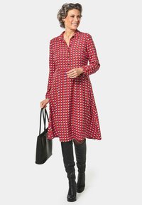Abito rosso con motivo geometrico, maniche lunghe e chiusura con bottoni, abbinato a stivali neri sopra il ginocchio e una borsa nera.