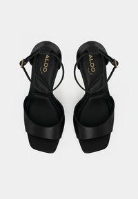 Mustat korkeakorkoiset sandaalit, joissa sileä pinta, leveä hihna varpaiden kohdalla, ohut nilkkahihna ja kultainen solkisomiste.